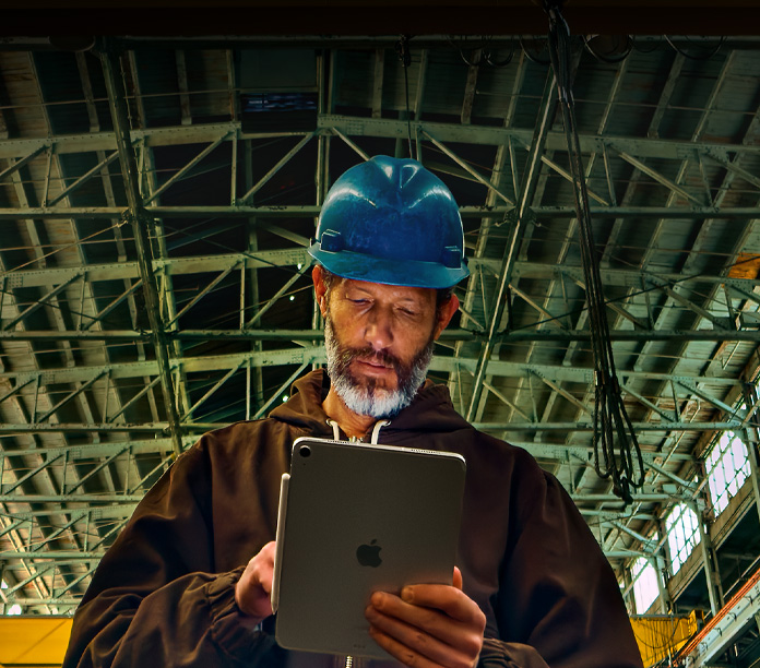 iPad, warna perak, dengan Apple pencil yang terpasang, dipegang oleh seseorang yang mengenakan jaket cokelat dan helm pelindung berwarna biru, sedang bekerja di lingkungan industri dengan balok-balok logam di atas kepala