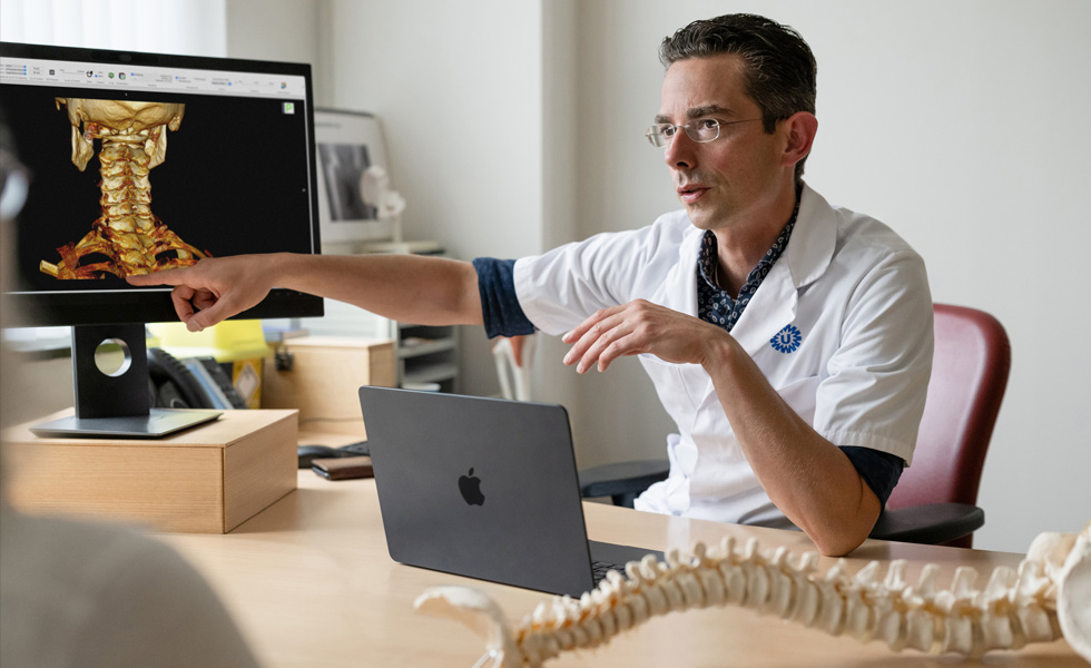 Un professionnel de la santé montre un rendu squelettique 3D de l’arrière d’un cou sur un Studio Display, avec un MacBook sur son bureau