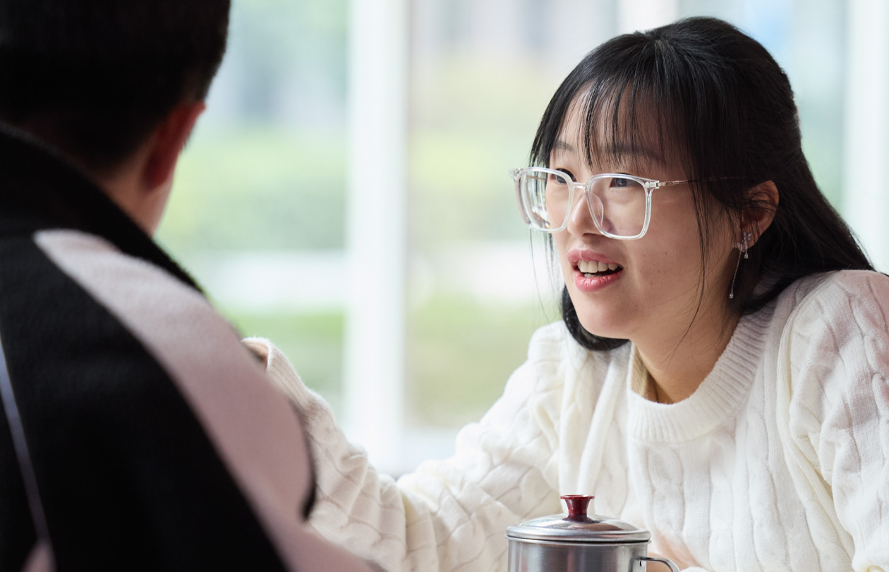 Two people sit facing each other at a cafeteria table and talk